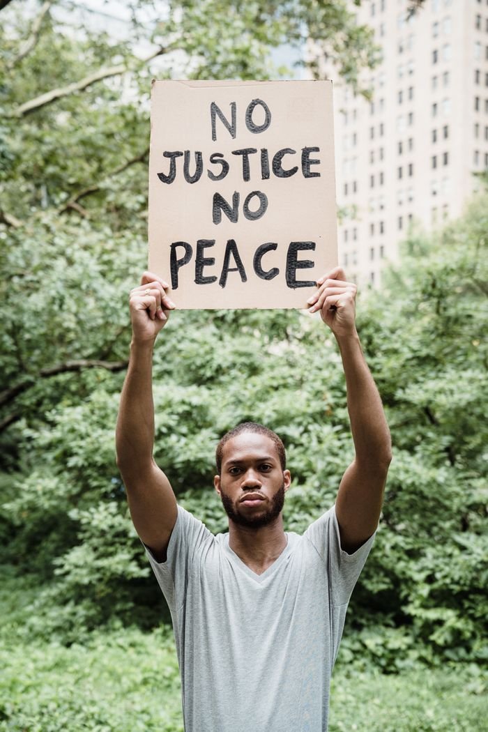 A man holding a 'No Justice No Peace' sign during a protest in a park setting.